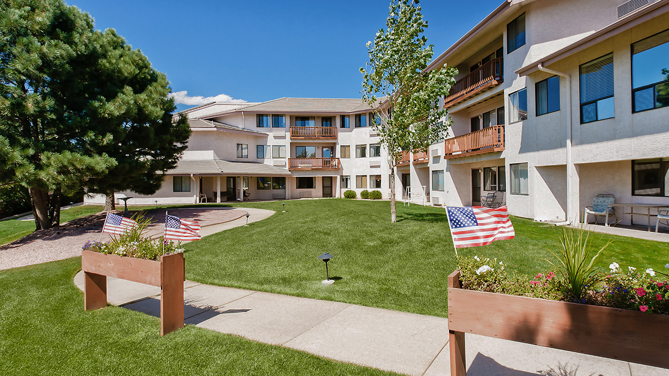 Exterior view of Holiday Sunridge retirement community in Colorado Springs with walking paths, patriotic flags, lush green space and patios.
