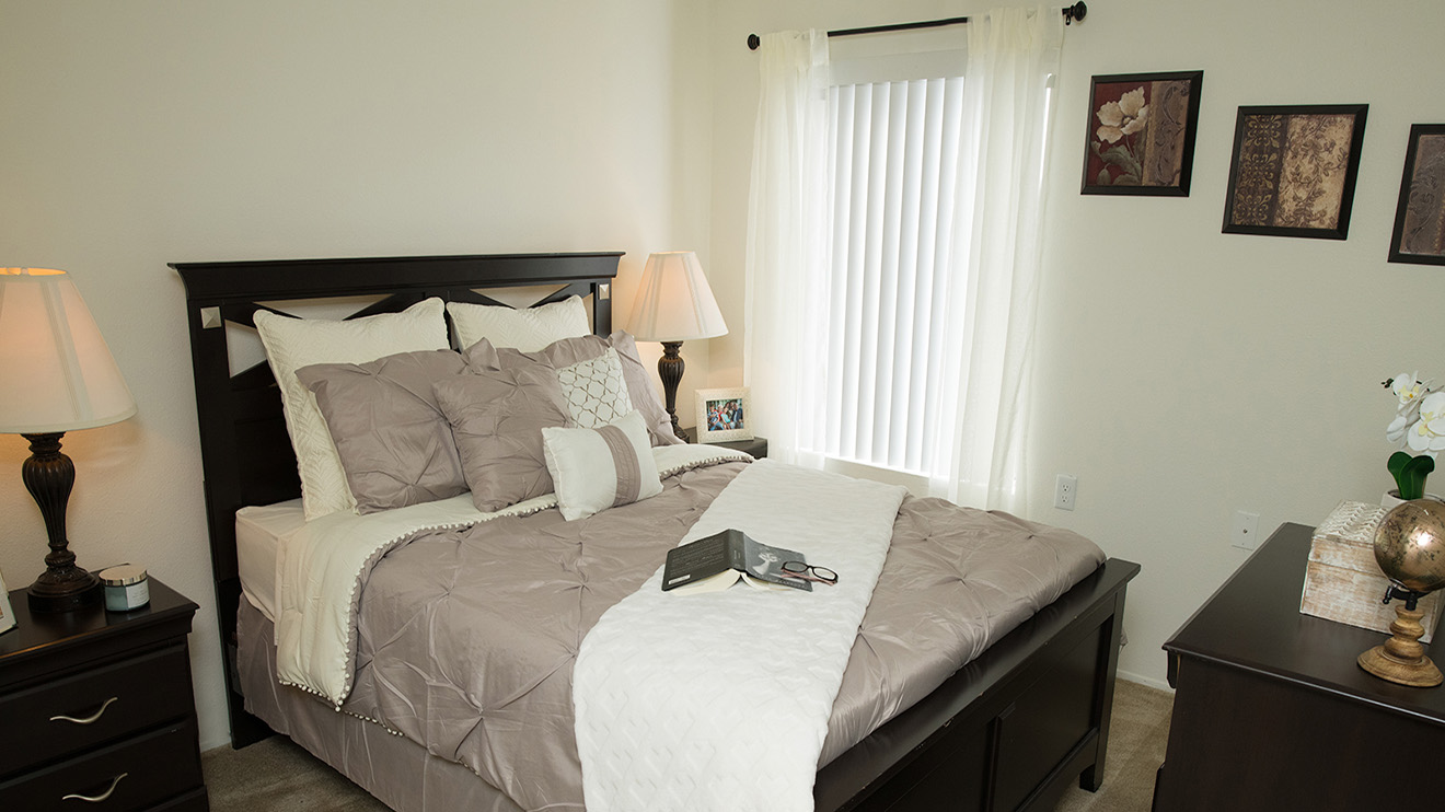 Senior apartment bedroom in Escondido, CA with open book on the bed, a dresser, pictures and curtains over a window.